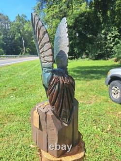 Colibri en bois sculpté à la tronçonneuse unique, en vol stationnaire au-dessus d’une fleur bleue